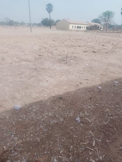 The image depicts an outdoor scene in Kanwa, Nigeria, under a hazy, light gray sky. The foreground consists of dry, reddish-brown earth extensively covered with debris, including plastic bags, white paper fragments, and small pieces of wood. A distinct, dark shadow cuts diagonally across the lower center of the frame. The midground features a wide, open expanse of barren, light-colored dirt with sparse, dry plant growth. In the background, a light-beige, single-story building with a light-colored roof and five rectangular windows is prominently situated on the right side, with a second, darker building partially visible behind it. To the left, a rudimentary metal goalpost stands on a dusty field, where several small, indistinct human figures are present. A tall, slender palm-like tree rises in the mid-background, positioned near a dark, thin vertical pole on the left, and a broader, leafy green tree is visible near the building on the right.