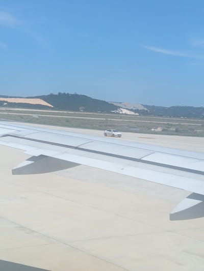 This image, taken from an airplane, shows a white car driving on a tarmac near the airport in Bắc Cam Ranh, Vietnam. The airport's runway and surrounding landscape, characterized by green vegetation and a distant hill with a sand pile, are visible. The airplane's wing dominates the foreground, with its reflective surface catching the sunlight. The sky is clear blue.