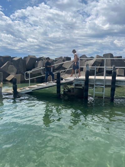 Two people are on a structure made of concrete blocks and metal railings, possibly a pier or breakwater. One person is standing at the edge looking over the railing, while the other appears to be climbing down a ladder into the water. The water is light green. The sky above is covered with puffy white clouds. The structure appears to be next to the ocean.