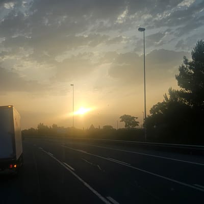 The image depicts a roadside scene at sunset or sunrise, with the sun low on the horizon and casting a warm, golden light. A truck, with its brake lights illuminated, is visible on the left side of the frame, partially obscuring the foreground. The truck appears to be traveling on a multi-lane highway. On the right side of the highway, a line of trees and vegetation is silhouetted against the bright sky. Two tall lampposts are present along the road, one closer to the foreground and another further in the distance. The sky is filled with scattered clouds, which are illuminated by the sun. In the distance, beyond the highway, there is a hazy landscape with what appear to be buildings and more trees. The scene is captured from a low vantage point, suggesting the photographer is likely in another vehicle on the same road. The overall atmosphere is serene and evokes a sense of travel. This scene is consistent with the landscape found along the AP-7 highway passing through or near Banyeres del Penedès, Spain, a region known for its vineyards and Mediterranean climate, which would explain the presence of trees and the characteristic golden light of sunsets in such locations.