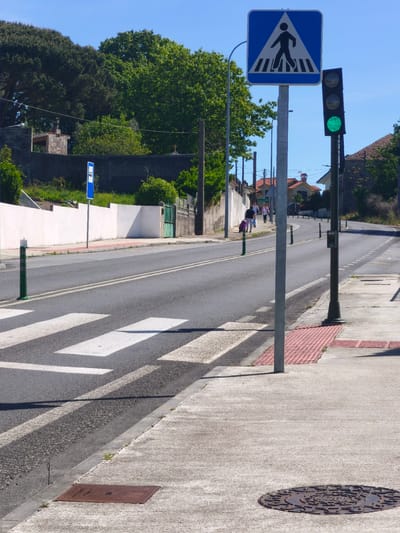 The image shows a street scene in Cangas de Morrazo, Spain. The street has a pedestrian crossing with white stripes and a blue pedestrian crossing sign. A traffic light is visible, displaying a green signal. On the sidewalk, there are two utility covers: one rectangular and one circular. The street ascends slightly, with green bollards positioned along the side. Several people are walking in the distance. A white wall runs along the left side of the street, with trees and greenery behind it. The sky is clear and blue.