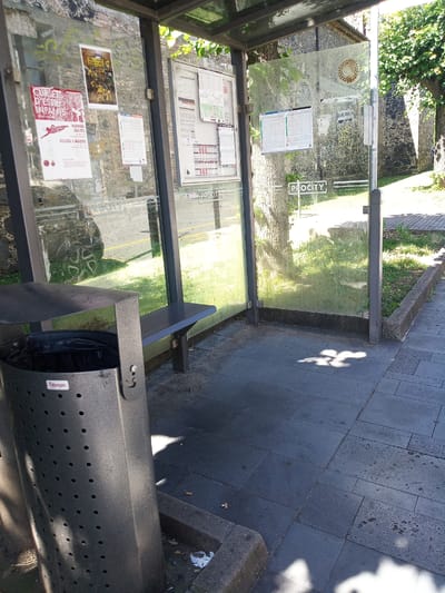The image shows a bus stop shelter in Castellfollit de la Roca, Spain, on a sunny day. The shelter has glass walls and a metal frame, with a metal bench inside. A black trash can with a "Fabregas" logo is positioned to the left of the bench. Posters are displayed on the glass walls, including what appears to be a bus schedule and advertisements. The ground outside the shelter is paved with dark tiles, and there is a grassy area beyond the pavement. Shadows of leaves and branches are cast on the pavement and the shelter walls, indicating the presence of trees nearby. The overall scene depicts a typical urban bus stop environment.