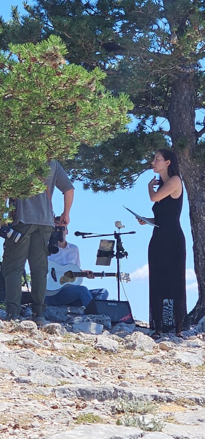 This image depicts an outdoor event taking place on a rocky, arid terrain with sparse vegetation under a clear blue sky. Dominating the scene are large trees with dense green foliage providing shade. In the foreground, on the left, a person is partially visible, wearing a grey t-shirt and olive-green pants, with a camera bag attached to their belt and holding a camera. To the right, a woman in a long black dress stands holding a document, her hand near her face. In the middle ground, a man in a white shirt and blue jeans is seated, playing a white acoustic guitar. A microphone stand with a small decorative item on top is positioned between the guitarist and the woman. A black amplifier with a "BOSS" logo is visible near the guitarist's feet. The overall impression is of a small, informal performance or ceremony in a natural, somewhat rugged environment. The location is identified as being in Općina Bol, Croatia, which is known for its beautiful rocky coastlines and Mediterranean vegetation.