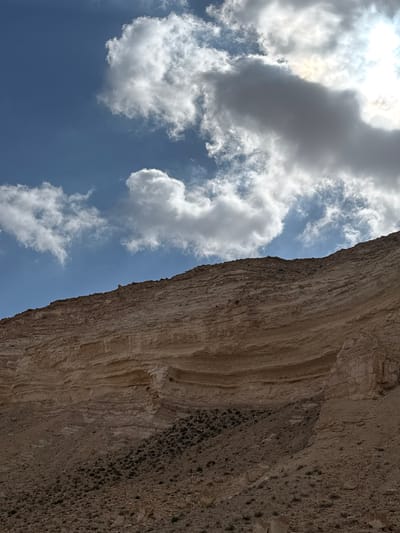 The image shows a view of a rocky cliff face in the Ramat Negev Regional Council, Israel, with a cloudy sky above. The cliff is composed of layered rock formations. The sky is blue with cumulus clouds. The image is rotated.