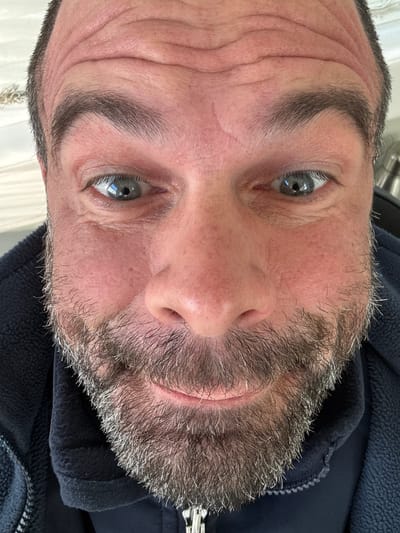 A close-up, slightly off-center, portrait of a middle-aged Caucasian man's face, oriented as if the viewer is looking up at him. He has graying hair and a salt-and-pepper beard. His skin is fair with visible wrinkles on the forehead. His eyes are gray. He is wearing a dark blue fleece jacket. There is no indication of the location being Diekirch, Luxembourg, or any other specific place. The background is indistinct.
