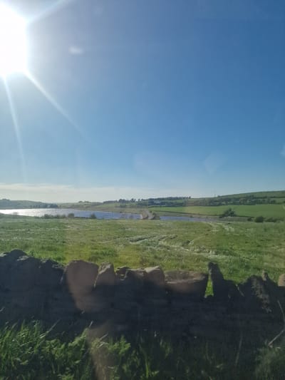 The image displays a wide, sunny landscape under a clear blue sky, viewed from a slightly elevated perspective. The main subject is a pastoral scene featuring vast green fields, a body of water, and a bright sun.
In the upper left portion of the image (when rotated correctly), a brilliant, star-shaped sun flare dominates, indicating a bright, clear day. The sky is a gradient of light to medium blue, with no clouds visible.
The midground features a long, narrow body of water, likely a river or canal, reflecting the sunlight in shimmering streaks. On either side of the water, expansive green fields stretch towards the horizon. The fields are a mix of light and darker greens, suggesting varied vegetation or cultivation. In the far distance, beyond the water and fields, more rolling green hills or fields are visible, extending to a faint horizon line. There might be some distant structures or trees, but they are not clearly discernible due to distance and light conditions.
The foreground of the image consists of a dark, rocky or earthy embankment, which appears to be on the right side of the water body. This embankment is partially covered with patches of grass and shows some irregular rock formations or large clods of earth, creating a textured contrast with the smooth fields and water. The overall impression is one of a tranquil, open-air environment, likely a rural area within Hyndburn, United Kingdom. No human activity or visible text is present.
