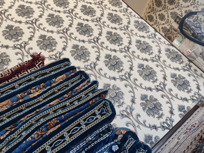 In Gurlan, Uzbekistan, an indoor scene shows rolled up rugs with intricate patterns against a wallpapered wall adorned with floral motifs. The rugs are layered and appear to be made of dark blue velvet with contrasting gold and white trim, and some feature maroon tassels. The wallpaper is a pale cream color with a repeating damask-like pattern in shades of blue and gray. A small portion of another rug is visible in the lower right corner, with a lighter color and red accents. The lighting suggests an indoor environment, possibly a room.