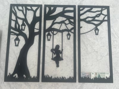 A triptych wall art made of black metal is displayed against a textured light-colored wall. The artwork depicts a stylized tree with lanterns hanging from its branches. The left panel features the trunk and lower branches of the tree with two lanterns. The middle panel shows a silhouette of a girl on a swing, suspended from a branch, with two lanterns visible. The right panel displays more branches with one lantern. At the bottom of the right panel, two Russian 5-ruble banknotes are partially visible, tucked into the decorative grass at the base of the artwork. The environment appears to be indoors, likely a room in Kokhma, Russia, given the currency displayed.
