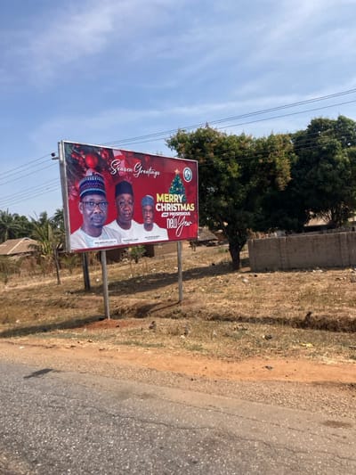 The scene, located in Ninzo West, Nigeria, depicts a roadside environment under a clear blue sky with scattered clouds. A prominent billboard stands in the mid-ground, displaying a red-themed "Season Greetings" message, "Merry Christmas and Prosperous New Year," and photographic images of three men identified as Dr. Abdullahi A. Bago, Dr. Babangida A. Gbazi, and Mr. Joshua M. Joji. The billboard is mounted on two vertical metal posts. In the immediate foreground, a section of a grey asphalt road with visible cracks runs horizontally. Adjacent to the road, the ground consists of dry, reddish-brown earth with sparse, desiccated vegetation and scattered refuse. In the background, to the left, are several low-lying structures with brown roofs and a cluster of palm trees. To the right, dense green trees and a partially constructed concrete block wall are visible. Overhead utility lines traverse the upper portion of the frame.
