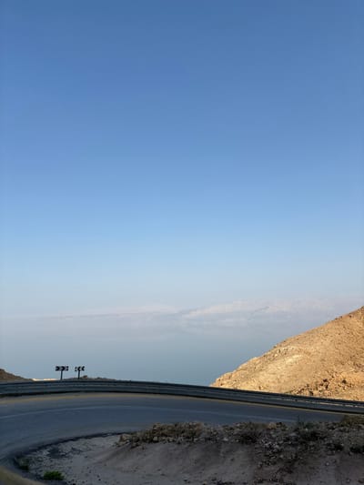 An elevated landscape photograph captures a winding asphalt road, featuring a sharp left-hand curve, in the Megilot Yam HaMelakh Regional Council, Palestinian Territory. The road is dark grey with faint white and yellow markings, bordered by a silver metal guardrail. Two black and white chevron road signs, indicating a sharp left turn, are positioned beyond the guardrail. To the right, a barren, rocky, tan-brown hillside descends. In the middle ground, a large body of light blue water, consistent with the Dead Sea, stretches into the distance, with hazy, light-colored mountains visible on the far horizon. The upper two-thirds of the image depict a clear, bright blue sky. No individuals, vehicles, or specific actions are present in the scene.