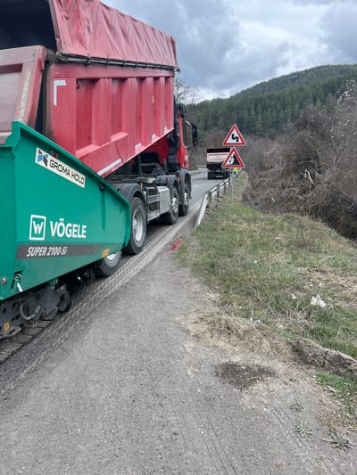 The image depicts a road construction or maintenance scene in Dagonovo, Bulgaria, under an overcast sky. In the foreground, a green Vögele SUPER 2100-5i asphalt paver is positioned on the left edge of an asphalt road, its textured tracks visible on what appears to be a newly laid or prepared surface. Adjacent to and slightly behind the paver is a red multi-axle dump truck, prominently displaying the "GROMA HOLD" logo, with its bed partially raised. A distinct red marking is present on the asphalt near the paver's tracks. A metal guardrail runs along the right side of the road, bordering a grassy, unkempt verge littered with dry vegetation and some white debris. Two triangular warning signs are mounted on a single post near the guardrail: the upper sign indicates a winding road, and the lower sign warns of falling rocks. Further down the road, a second dump truck is visible in the distance. The background features a dense, coniferous forest covering a hillside. No individuals are visible in the scene.