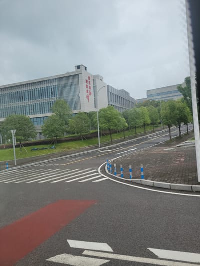 The image shows a street scene in Yufengshan, China. In the background, there is a modern, multi-story building with a glass facade and some red Chinese characters on its side. In front of the building, there is a row of green trees and manicured grass. A road with pedestrian crossings runs through the foreground. There are also several blue and white bollards lining the edge of the road. The sky is overcast.