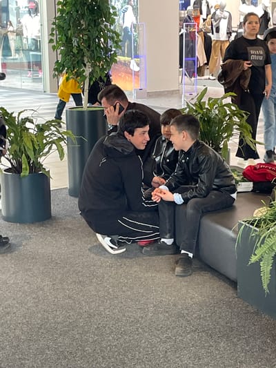 In a brightly lit interior, possibly a shopping mall or commercial space in Саларьево, Russia, a group of people is gathered around seating areas adorned with potted plants. Three individuals are situated on a dark gray sofa; two are interacting closely, while another sits nearby. One person is wearing a black leather jacket and dark pants, while another wears a black hooded jacket with white stripes on the sleeves and dark pants. Shoes of varying colors are visible on their feet. The environment includes retail displays with mannequins and clothing racks in the background, as well as additional individuals. The floor is covered with a textured gray carpet.