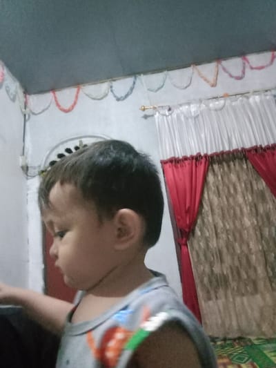 The image captures a young child, approximately a toddler, in an indoor setting in Lam Ujong, Indonesia. The child is seen in profile, facing towards the left side of the frame, from the chest up. The child has dark hair and is wearing a light gray sleeveless top with colorful designs. The child's left arm is slightly raised. The background consists of light-colored walls, a darker gray ceiling, and a colorful patterned mat on the floor featuring geometric shapes. Along the upper wall, decorative scalloped garlands in various light colors are strung. To the right, a window is partially visible, adorned with white sheer curtains layered over patterned brown curtains, tied back with red fabric, all suspended from a gold-colored curtain rod. On the left wall, a vertical electrical conduit and an arched decorative element with dark detailing are visible. The scene is illuminated by ambient light.