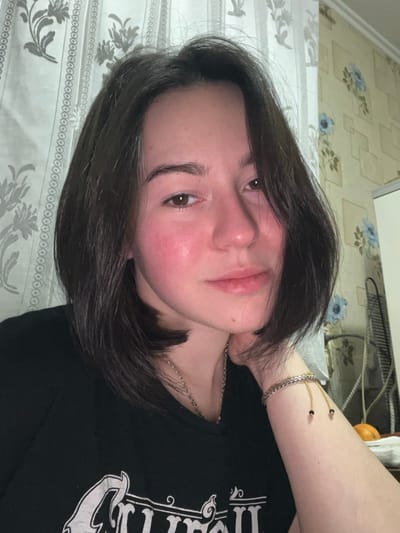 A woman with short, dark hair and fair skin is looking at the camera. She has a chain necklace, a silver bracelet, and a corded bracelet with dark beads. She is wearing a black t-shirt. The background includes a bed with a patterned bedspread and a wall with floral wallpaper.