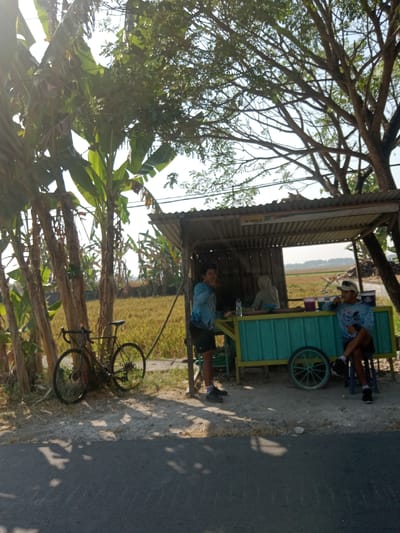 The image depicts a scene in Kemamang, Indonesia, on a bright, sunny day, with dappled sunlight filtering through trees. In the foreground, a paved road is visible, with shadows cast upon it. To the left, a black bicycle is leaning against several banana trees. Behind the trees and bicycle is a field of golden-yellow crops, likely rice.
Dominating the mid-ground is a small, open-air stall with a corrugated metal roof supported by wooden posts. A teal-colored cart with yellow trim and a prominent wheel is positioned in front of the stall. Inside the stall, two individuals are present. One person, wearing a light blue shirt and dark shorts, is standing and appears to be gesturing. Another person, wearing a grey hooded garment, is seated behind a counter. To the right, another person, wearing a light blue shirt, a cap, and dark shorts, is seated on a blue stool, with their legs crossed. They appear to be eating or holding something.
The environment suggests a rural or semi-rural setting, possibly a roadside rest stop in an agricultural area. The overall atmosphere is casual and relaxed.