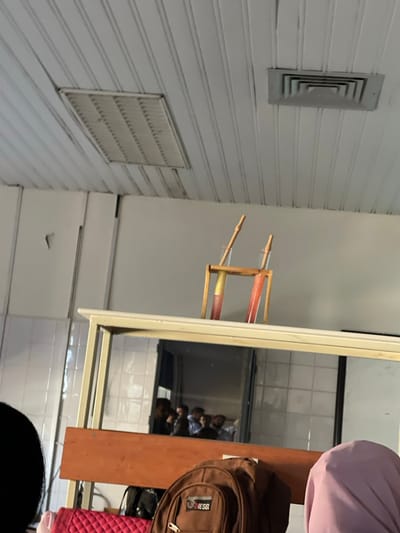 The scene is an indoor environment in قرية الضبية, Syria, characterized by a white paneled ceiling with visible wear, featuring a rectangular louvered light fixture frame on the left and a square air vent on the right. Below the ceiling, a light-colored wall shows minor imperfections. In the mid-ground, a light-framed table or counter with a brown horizontal surface is present. On this surface, two clear cylindrical glasses are displayed in a small wooden stand. Each glass contains a layered yellow and red liquid, with a wooden straw inserted. Below this counter, a reflection in a window or mirror reveals a group of indistinct individuals, likely men, standing in an adjacent tiled area. In the foreground, partially visible, are a brown "DIESEL" backpack, a red quilted bag, a person with dark hair on the left, and a person wearing a light purple hijab on the right.