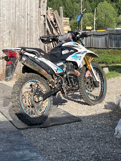 The media file shows a dirt bike parked outdoors. The motorcycle is white with blue and black accents and has knobby tires, suggesting it is designed for off-road use. It is leaning on its kickstand. The bike appears dirty, with mud splattered on the tires and fenders, indicating it has recently been ridden.
The setting appears to be a rural or semi-rural area, possibly a backyard or a workshop. Behind the motorcycle, there is a wooden fence and some stacked lumber. Green foliage and trees are visible in the background. The ground is covered with gravel and a dark, textured surface, possibly a tarp or rubber mat. Sunlight is casting shadows, suggesting it is daytime, likely a sunny day.
There is no visible activity or event taking place; the motorcycle is stationary. Notable details include the design of the motorcycle, its muddy condition, and the rustic outdoor setting.
Visible text on the motorcycle includes the brand name "CFA" on the exhaust pipe and some decals on the fairing. The text is in English.
The location context is Minyar, Russia.