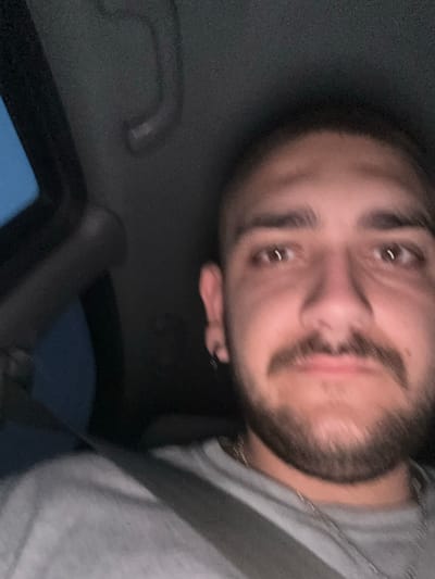 A close-up, selfie-style photograph captures a young man inside a vehicle. He has short, dark hair and a mustache and beard. He is wearing a grey t-shirt and a silver chain. The man is looking directly at the camera. The interior of the vehicle is dark, with a sunroof visible at the top left of the frame. The image appears to have been taken at night. There is no visible indication of the specific location within Staro Selo, Bulgaria.
