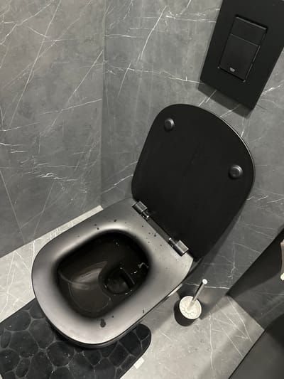 The image shows a top-down view of a black toilet in a bathroom. The toilet lid is partially open. Next to the toilet is a black flush plate and a toilet brush. The bathroom floor is tiled with a gray marble pattern. A black bath mat is partially visible to the right of the toilet.