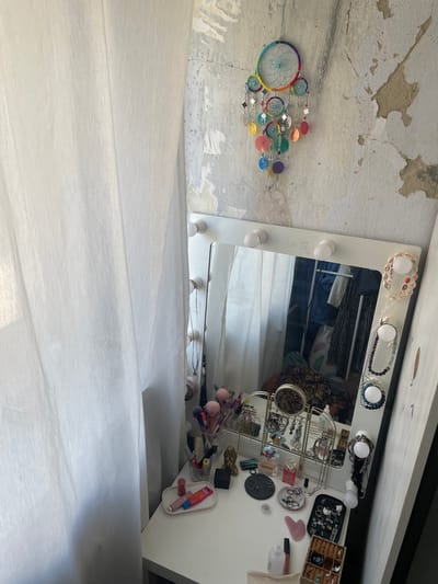 The image depicts a dressing table area with a vanity mirror illuminated by light bulbs, positioned against a distressed, textured wall. To the left, a sheer white curtain drapes, obscuring a window. On the table, various makeup items are arranged, including lipsticks, brushes in a container, and a makeup palette. Several pieces of jewelry, such as necklaces and rings, are displayed on organizers and scattered across the surface. A dreamcatcher with colorful elements hangs on the wall above the mirror. The overall scene suggests a personal grooming and beauty space. The provided information about the location in Kopeysk, Russia, is not visually verifiable from the image itself.