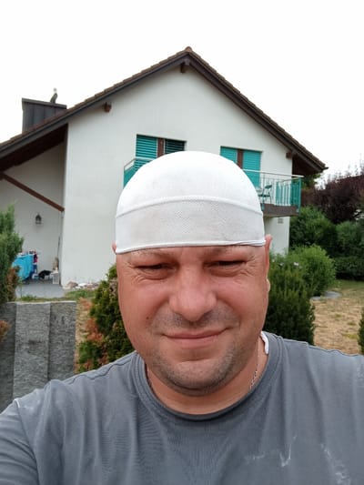 A man wearing a white skullcap and a grey t-shirt takes a selfie outdoors. He has a light beard and is smiling slightly. Behind him, a white house with a dark brown roof and green shutters is visible. A balcony with a green railing juts out from the upper story of the house. Various bushes and trees surround the house, and a grey stone structure is in the foreground to the left. The scene appears to be in a residential area.