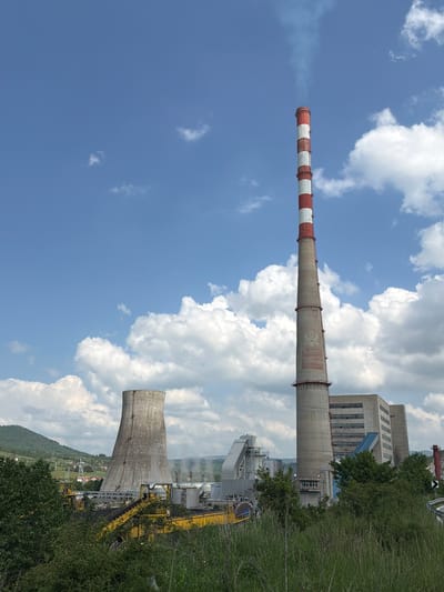 The image shows a coal-fired power plant located in Zabrđe, Montenegro. A tall smokestack with red and white bands is visible against a blue sky with puffy clouds. Adjacent to the smokestack is a large, gray industrial building. Nearby, there is a cooling tower, also gray. Yellow machinery and conveyor belts are positioned near the cooling tower. Trees and vegetation are visible in the foreground and surrounding the plant. In the distance, a small village or town is nestled against a mountain.