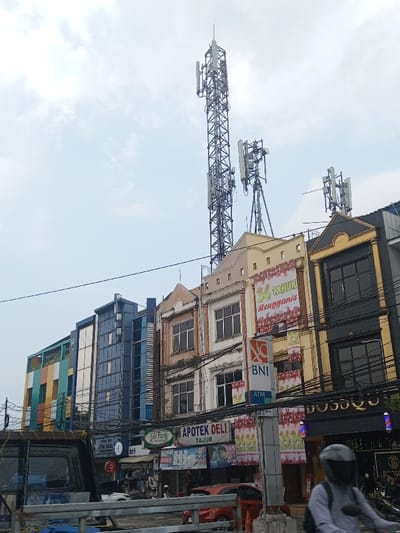 The image captures a vibrant urban street scene, likely a commercial district, under a bright, partly cloudy sky. The main focus is on a row of multi-story buildings that line the street, topped with numerous communication towers and antennas.
The sky occupies the upper left portion of the image, appearing light blue with scattered white clouds, suggesting a daytime shot, possibly under an overcast or hazy condition.
Prominently featured above the buildings are three distinct communication towers. The tallest and most complex one is a metal lattice structure, heavily equipped with multiple rectangular antenna panels at various heights, indicating a significant telecommunications hub. To its right, two smaller, yet still substantial, antenna arrays are visible on different rooftops, each consisting of poles with several antenna panels attached.
The buildings themselves display a mix of architectural styles and colors. From left to right:
- The leftmost building is modern, featuring a distinctive facade with large, colorful panels in green, blue, yellow, and orange.
- Adjacent to it is a taller, dark blue building with a more contemporary, grid-like facade, possibly made of glass or metal.
- Following that, a beige-colored building with a more classic design, characterized by arched window frames on the upper floors and some visible foliage growing on its exterior, suggesting age.
- Next is a light yellow building, similar in style to the beige one, which features a large banner draped across its upper floors.
- The rightmost visible building is dark brown or black, sharing a similar traditional architectural aesthetic with the beige and yellow buildings, complete with dark-framed windows.
At street level, the scene is bustling with activity. The front part of a dark-colored truck, possibly a pickup or utility vehicle, is visible on the far left. Numerous utility lines, including power and communication cables, crisscross above the street and in front of the buildings.
Several commercial signs are visible on the lower facades of the buildings:
- On the yellow building, a large banner proclaims "34 TAHUN Reuggansi," with smaller banners below it reiterating "34 TAHUN." Below this, a prominent vertical sign displays the "BNI" logo with "ATM" underneath, indicating a bank and an ATM facility.
- Below the beige building, a sign reads "APOTEK DELI TAJUR," identifying a pharmacy.
- Further left, partially obscured signs include "Ide Paris" and "bft."
- On the dark brown building to the right, a sign with "BO$$QU" is visible.
People and vehicles contribute to the lively street atmosphere. On the far right, a person wearing a helmet is riding a motorcycle, moving away from the camera. Other indistinct vehicles and people are visible further down the street, suggesting a moderately busy urban thoroughfare. The overall impression is that of a typical commercial street in a densely populated area.