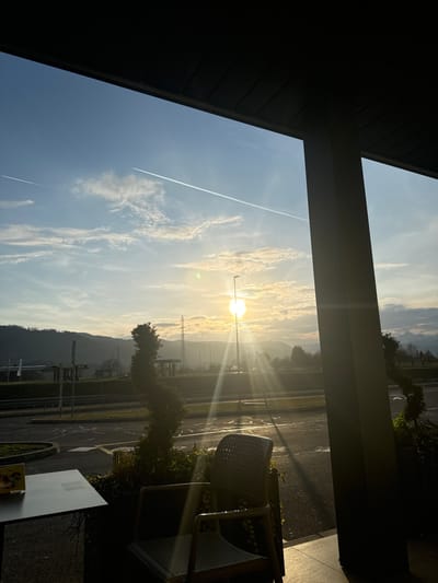 The image depicts an outdoor scene observed from a covered patio or terrace, located in Radovljica, Slovenia. The perspective is outward, beneath a dark, horizontally ribbed overhang, with a thick, light-colored support pillar visible on the right side of the frame.

The foreground features a partial view of a dark, rectangular outdoor table on the left, displaying a yellow object, possibly a menu or leaflet. To its right, a light-colored woven outdoor chair is positioned, facing the distant landscape. Between the seating area and the midground, several manicured shrubs or small trees, contained in planters, are visible. One prominent shrub in the center foreground exhibits a distinct spiral or twisted form.

The midground consists of a paved surface, indicative of a parking lot or a section of a road, with faint white markings. Infrastructure such as a light pole with a bright star-shaped sun flare directly behind it, and power lines/towers, are discernible.

The background reveals a range of hills or mountains under a partly cloudy sky. The sun is low on the horizon, directly in the center-right of the frame, emitting strong light rays and lens flare, backlighting the scene and creating a hazy atmosphere over the distant landscape. Two distinct contrails from aircraft are present in the clear blue sky, one extending diagonally from the top-right to the mid-left, and another shorter one in the far upper-left. No people, animals, or specific actions are depicted. The overall lighting suggests either sunrise or sunset.