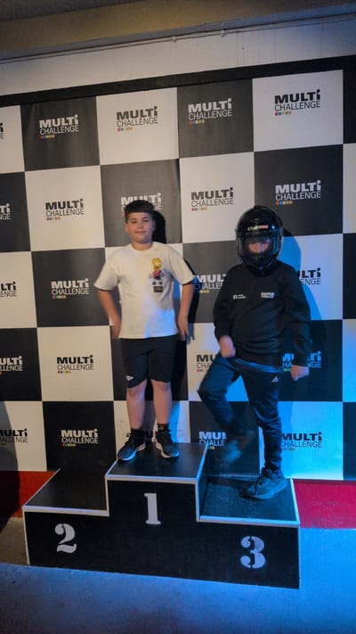 This image depicts two young individuals, likely boys, participating in an activity at what appears to be an indoor entertainment or competitive venue. The setting features a checkered black and white backdrop with the repeated text "MULTI CHALLENGE" displayed prominently. One boy is dressed in a black hooded sweatshirt and a black helmet, with his body in a dynamic pose, suggesting movement or an action shot. He is standing on a raised podium with the number "3" visible. The second boy, wearing a white t-shirt with a graphic print and black shorts, is positioned on a lower step of the podium, marked with the number "1". His posture is more relaxed, and he is looking towards the camera with a slight smile. The podium itself is black with white numbers indicating different positions. The lighting appears to be artificial, with a blue hue in the background, characteristic of an indoor event space. The overall atmosphere suggests a competitive or celebratory moment, possibly related to go-karting or a similar racing-themed activity, given the helmet and the podium. The visible text "MULTI CHALLENGE" reinforces the theme of competition. The image is a vertical orientation.