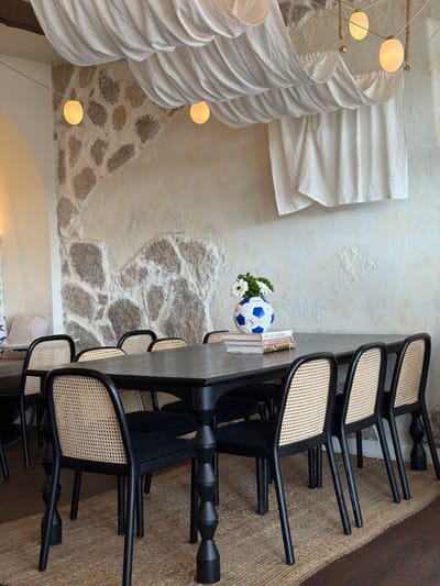 The image depicts a dining area with a rustic and modern aesthetic. The scene is dominated by a long, dark wooden table surrounded by black chairs with woven seats. On the table, a blue and white soccer ball sits next to a small bouquet of white flowers and a few books. The background features a textured white wall with exposed stone elements and large, draped white fabric installations that resemble lampshades. Several light fixtures, with round, glowing bulbs, hang from above. The floor is covered with a natural fiber rug. The lighting suggests it might be daytime or evening, with the warm glow of the lights creating an inviting atmosphere. The overall impression is one of casual elegance, possibly in a restaurant or a well-decorated home. There is no visible text in the image.