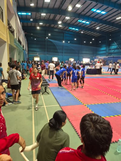 This media file depicts an indoor sports event, likely a kabaddi match or tournament, taking place in Phagwara, India. The scene is a large gymnasium with a high ceiling and a distinctly marked court, divided into red and blue sections. Rows of people, seemingly participants and spectators, are visible on the court and along the sides. Many individuals are wearing athletic attire, predominantly blue and red.
In the foreground, two individuals are captured from the back, one wearing a red hooded sweatshirt and the other a green athletic top. Their presence suggests they are part of the event, either as participants waiting for their turn or as observers. The general atmosphere appears to be one of an organized sporting event, with participants lined up and ready.
The background shows the structure of the gymnasium, with large windows and lighting fixtures on the ceiling. The presence of a large net and other sports equipment is also implied by the setting. The red and blue mats on the court, along with the white lines marking the boundaries, are characteristic of kabaddi courts.
The image is taken during the daytime, evidenced by the natural light filtering through the windows. The focus is primarily on the people and the sports arena, with no specific indication of weather conditions. The overall mood seems to be energetic and competitive, typical of a sporting event. No discernible text is visible in the image, other than what might be on clothing or signage in the distance, which is illegible.