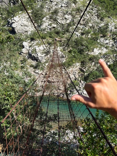 The image depicts a steep, rocky canyon in an unknown location within Montenegro, viewed from a high vantage point overlooking a deep chasm. A dilapidated suspension footbridge, constructed from rusted metal mesh and thick steel cables, stretches diagonally from the lower left foreground to the mid-ground, spanning a body of clear, turquoise-green water. The riverbed is visible beneath the translucent water, appearing light-colored and stony. The canyon walls consist of rugged, light grey rock formations, partially covered by sparse green scrub and small trees. Two diminutive human figures are discernible on the far end of the bridge, positioned on the opposing bank of the canyon. In the lower right foreground, a blurred, light-skinned human hand, exhibiting tan lines, is partially visible, with the thumb and index finger extended in a gesture that appears to frame or point into the scene. The entire scene is brightly illuminated by direct sunlight, suggesting clear, fair weather.