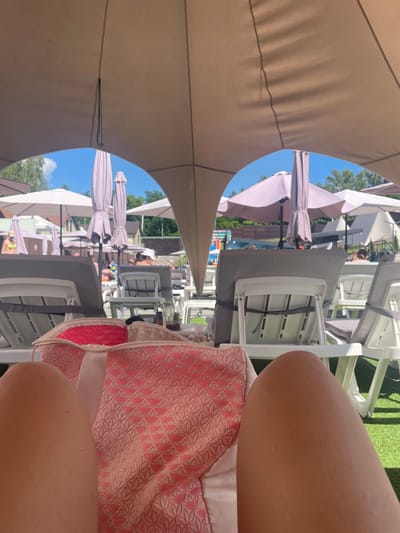 The image depicts a first-person perspective at a sunny outdoor swimming pool area in Zazymia, Ukraine. The foreground is dominated by a pink and white patterned beach bag and the bare legs of the photographer. In the midground, rows of empty, grey-cushioned lounge chairs are set up on a green artificial turf surface. Behind the chairs, a variety of light-colored umbrellas provide shade, with several closed and a few open. Beyond the umbrellas, there are trees and buildings, suggesting a resort or recreational facility. The sky is clear and blue with some scattered clouds. Several people can be seen in the background, some relaxing on chairs, others near the pool or in a shallow water area. The overall impression is one of a leisurely summer day at a poolside resort.
