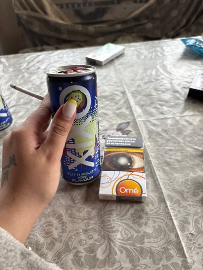 An indoor scene in Kastel, Bulgaria, presents a close-up view of a human hand holding a beverage can with a cigarette, positioned on a patterned tablecloth. The hand, displaying manicured nails with yellow tips and a faded tattoo on its dorsal surface, grips a blue and green can labeled "OKIX TUTTI FRUITY KIWI FLAVOUR." A lit cigarette, featuring a visible ash tip, is inserted into the can's opened drinking aperture. To the right of the can rests a white and orange "Omé" brand cigarette pack. The pack displays a Bulgarian health warning, including text that translates to "SMOKING INCREASES THE RISK OF BLINDNESS," accompanied by a graphic image of a severely diseased human eye. The tablecloth exhibits a light-colored, repeating floral or damask pattern. The background is blurred, showing a dark, patterned fabric object and indistinct lighter-colored items on a surface behind the main subjects.