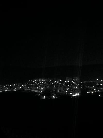 This monochromatic, high-angle, long-distance photograph captures the city of Bornarevo, Bulgaria, during nighttime. The urban area is illuminated by numerous artificial light sources, appearing as distinct white points against the dark backdrop of the landscape and sky. The lights delineate buildings and infrastructure, suggesting a sprawling settlement across varied terrain. No specific structures are clearly identifiable due to distance and low light conditions. The foreground is obscured by deep darkness, providing no discernible details. There are no individuals, vehicles, or overt actions visible within the frame. The environment is consistent with a populated urban or suburban area observed from an elevated or remote vantage point.