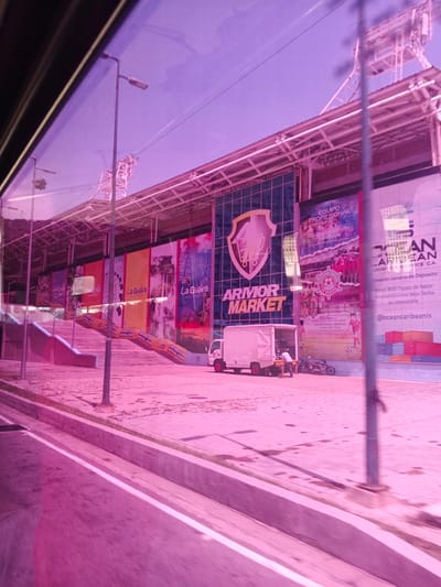 The image shows the exterior of a stadium or large building with a pinkish hue, suggesting it was taken at dusk or dawn, or that the image has been color-filtered. The building features large banners with graphics and text, some of which are partially obscured. One prominent banner displays the text "ARMOR MARKET" in large, bold letters. Another banner has the text "EQUIPO PROFESIONAL" above an image of a soccer team. There are also banners with the text "La Guaira" and what appears to be "APUESTAS ROYAL.COM".
In front of the building, there is a paved area with a sidewalk and a road. A white truck is parked in front of the building, with a person near it. A motorcycle is also visible. The scene appears to be outdoors, possibly in Macuto, Venezuela, given the context. The lighting is soft and diffused, contributing to the overall pinkish tone of the image. The overall impression is of a commercial or sporting venue, with advertising and branding prominently displayed. The scene appears calm, with no indication of a specific event or crowd.