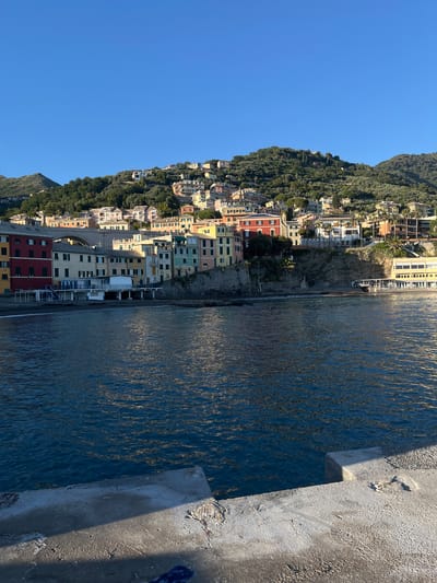 This image depicts a coastal view of Bogliasco, Italy. The scene shows colorful buildings nestled on a hillside adjacent to the Ligurian Sea. The buildings are a mix of residential and commercial structures, painted in various colors such as red, yellow, and pastel shades. Dense vegetation covers the upper part of the hillside. The sea is blue, with light reflecting off the water's surface. On the right side of the image, there is a concrete structure that appears to be part of a pier or walkway along the coast. The sky is clear and blue.
