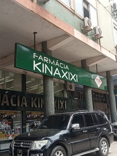 The image captures a street scene in Luanda, Angola, featuring the storefront of a pharmacy. A large, horizontal green sign prominently displays "FARMÁCIA KINAXIXI" in white capital letters. "FARMÁCIA" is Portuguese for "Pharmacy," and "Kinaxixi" is a known area in Luanda. A smaller red and white logo, possibly "MS," is also visible on the right side of the main sign.
The sign is suspended under a concrete overhang, which is supported by several cylindrical pillars seemingly clad in mosaic tiles. Below the main sign, the pharmacy's storefront windows show festive white decals, possibly snowflakes, along with partial text like "Festas" (Parties/Festivities) and "Feli" (likely part of "Feliz" - Happy). Another sign on the facade below the main one reiterates parts of "FARMÁCIA KINAXIXI" and includes the text "MONIZ SILVA IN".
A black Mitsubishi Pajero SUV is parked directly in front of the building, partially obscuring the lower facade. Its license plate, "LD 842 ZGP," includes "LD," a common prefix for Luanda vehicles. The multi-story building above the pharmacy has windows and several external air conditioning units mounted on its green, textured facade. The concrete elements of the building appear somewhat weathered. The scene is illuminated by bright daylight, suggesting it's either morning or afternoon, with no specific weather events discernible. No people are visible, and no specific activity is taking place; it is a static snapshot of a commercial street.