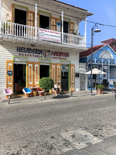The image depicts a street scene in Puerto Plata, Dominican Republic. In the foreground, a cobblestone street leads towards a building with a white facade and a balcony. The building houses "HELADERIA REPOSTERIA MARIPOSA AUSTRÍACA." Bright orange shutters and doors adorn the storefront. In front of the establishment, there are several uniquely shaped chairs resembling ice cream cones, and a small, round table. A young man is seated on one of the ice cream cone chairs, looking down. To the right of this building, a vibrant blue building with intricate white trim is visible, featuring a covered outdoor seating area with an umbrella and tables. A street lamp stands between the two buildings. The sky above is clear and blue. The environment suggests a sunny day in a lively urban area, likely a commercial district with unique architectural styles and ice cream shops.