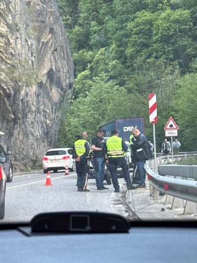 The image shows a roadside scene near Бачковски манастир, Bulgaria, with a group of people, some of whom appear to be law enforcement officers wearing neon vests. A white car is parked behind them, accompanied by orange cones. A large "DSV" truck is also present. The setting is alongside a road barrier with warning signs. The background features a rocky cliff face and a forested area. The image is taken from inside a vehicle, likely behind a window.