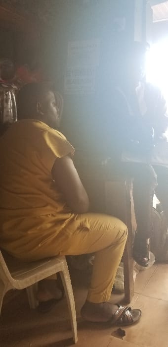 The image displays an indoor scene within a commercial establishment in Ado-Ekiti, Nigeria. Two adult males are seated at a wooden table, facing each other. The individual on the left, viewed from a partial rear-side profile, wears a loose-fitting, light brown or mustard-yellow short-sleeved top and matching trousers. He is seated on a light-colored plastic chair and wears dark-colored open-toed sandals. His right arm is visible, resting on the table. The individual on the right is heavily backlit by a bright light source, rendering him largely in silhouette. He appears to be wearing a dark jacket over a white collared shirt, with dark dress shoes visible beneath the table. His right hand rests on a document or papers on the table. The interaction suggests a conversation or a business transaction. On the wall behind the right individual, multiple white rectangular signs with dark lettering are visible, including phone numbers and "MONEYPOINT POS" (Point of Sale), indicating a financial or agent banking service. The floor is tiled in a light color. The environment is generally dim, with significant glare from the right side of the frame.
