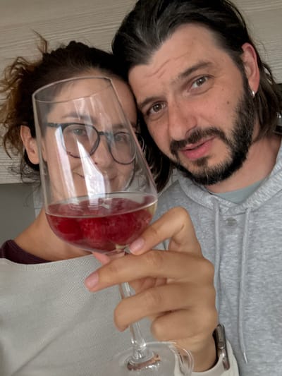 A man and a woman pose closely together with a glass of red wine in an indoor setting. The man has long, dark hair, a beard, and is wearing a grey hooded sweatshirt. The woman has glasses and is positioned behind the wine glass. The environment is nondescript and does not suggest a location in Ampfing, Germany.