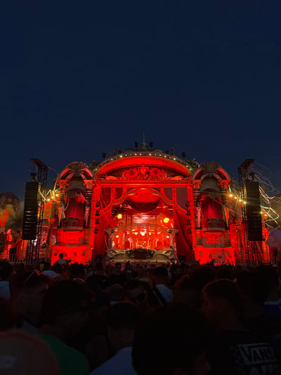 In Rumst, Belgium, at night, a crowd of people gathers in front of an elaborate stage bathed in red light. The stage, designed with ornate golden architectural details, draped red curtains, and various decorative elements, serves as the focal point. Large speaker stacks are positioned on either side of the stage, connected by scaffolding and adorned with streamers. The overall atmosphere suggests a lively outdoor event, possibly a music festival. The sky above is dark blue, indicating the time of day is after sunset.
