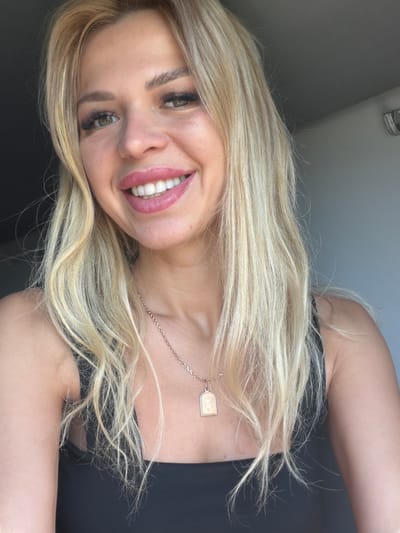 A medium shot shows a woman with blonde hair and fair skin smiling at the camera. She is wearing a black tank top and a rose gold necklace with a pendant. Her hair is long and wavy, falling around her shoulders. The background is blurry and appears to be indoors, with a grey wall and a dark area to the left. There is no indication of the location being Mykulychyn, Ukraine.