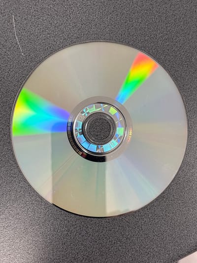 The image shows a compact disc (CD) lying on a textured gray surface. The CD is positioned centrally in the frame, with its reflective surface displaying a spectrum of rainbow colors due to light refraction. The edge of the CD is visible, and the center has a metallic hub with holographic patterns. A thin, white strand, possibly a hair or fiber, is present on the gray surface to the upper left of the CD. The surface appears to be a desk or table. There is no indication of the city of Mülheim an der Ruhr, Germany, in the image.