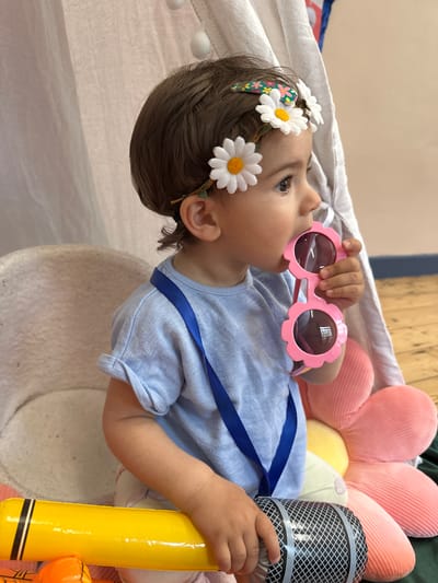 A young child, likely a toddler, lies on their back in what appears to be a play area. They are wearing a light blue t-shirt adorned with a dark blue ribbon. A daisy-themed headband is in their hair, and they are holding pink, flower-shaped sunglasses up to their face, seemingly about to put them on or examine them.
Behind the child, a large, plush pink flower pillow is visible, along with a yellow inflatable microphone. The setting includes a white, textured surface, possibly a mat or cushion, and a draped white fabric, hinting at a playful or cozy environment. The lighting suggests daytime. The child's gaze is directed upwards and to the left, with a curious or playful expression. The image is taken from a high-angle perspective. The overall mood is lighthearted and innocent. No visible text is present in the image.