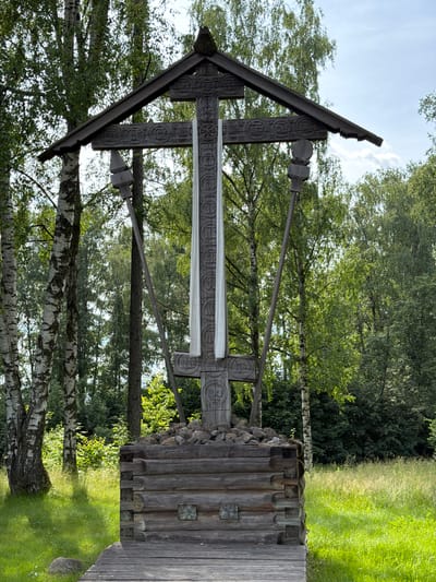 The image shows a large wooden cross structure with intricate carvings. The cross is adorned with a white cloth draped vertically down its center. The structure is supported by a base made of rough-hewn logs, topped with stones. It stands in a grassy area with trees in the background, including birch trees with distinctive white and black bark. The scene appears to be set in a park or natural environment, with a clear sky above. The location is identified as Nevskii Parkleskhoz, Russia. There are no people visible in the image.