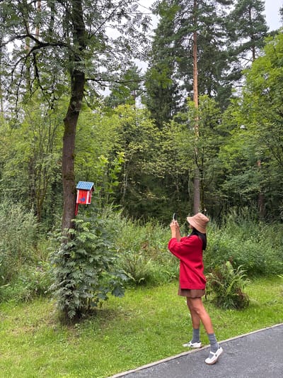In Pavlovskaya Sloboda, Russia, a person stands on a paved path in a park-like setting, facing away from the camera. They are wearing a light brown bucket hat, a red long-sleeved shirt, and a brown skirt. The person is holding a mobile phone in their hands, appearing to take a photo. The environment is lush with green foliage and tall trees. A red and blue birdhouse is mounted on a tree trunk to the left of the frame. The ground is covered in grass and a gravel path is visible in the foreground. The sky is overcast.