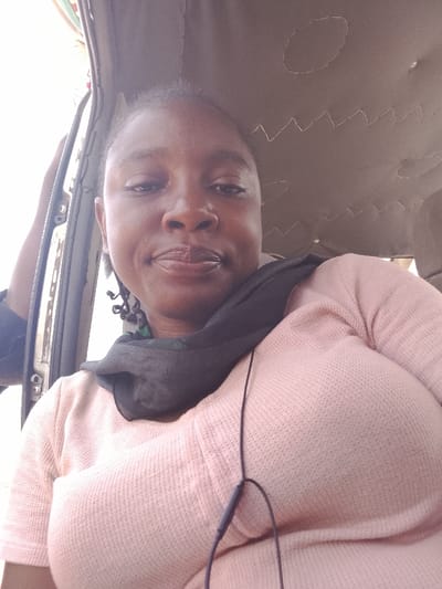 The image depicts an adult female situated within the interior of a vehicle. She has dark skin and short, dark, coiled hair, and is looking directly at the camera with a neutral to slightly smiling expression. She is wearing a light pink, long-sleeved, waffle-knit shirt and a dark scarf around her neck, which exhibits a subtle green pattern on its left side. A black wired earphone extends down her chest.
The vehicle's interior features a light-colored fabric headliner with decorative zigzag and oval stitching. To the subject's left, a portion of an open vehicle door frame or window is visible, through which bright exterior light is entering. A dark-sleeved arm is partially discernible on the far left, resting near the door frame.
There is no visual information within the image to ascertain the specific location as the city of Ibi, Nigeria, or any other precise geographical point. The scene is illuminated by ambient daylight.